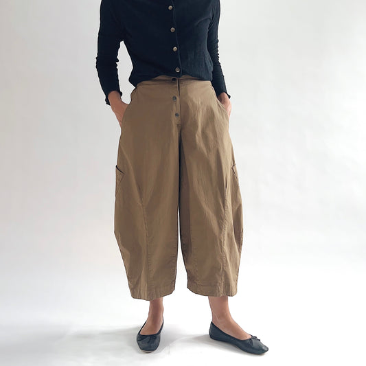 Full front view of model wearing Eleven Stitch winter olive snap front pant with hands in pockets, showing snap front waistband, wide balloon leg silhouette, large side patch pockets, gathered elastic ankle hem, and warm olive tan color, styled with black button front top and black ballet flats