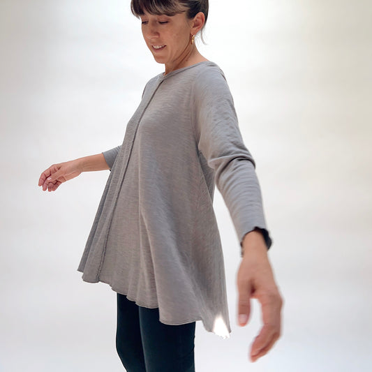 North Star Base Double Cotton Shorter Hi Low Front Seam Top in Zinc, side view on smiling model in motion, zinc gray double-layered cotton with raw hem crew neckline, visible front center seam, long sleeves with raw cuffs, and flared hi-low hem, paired with dark jeans
