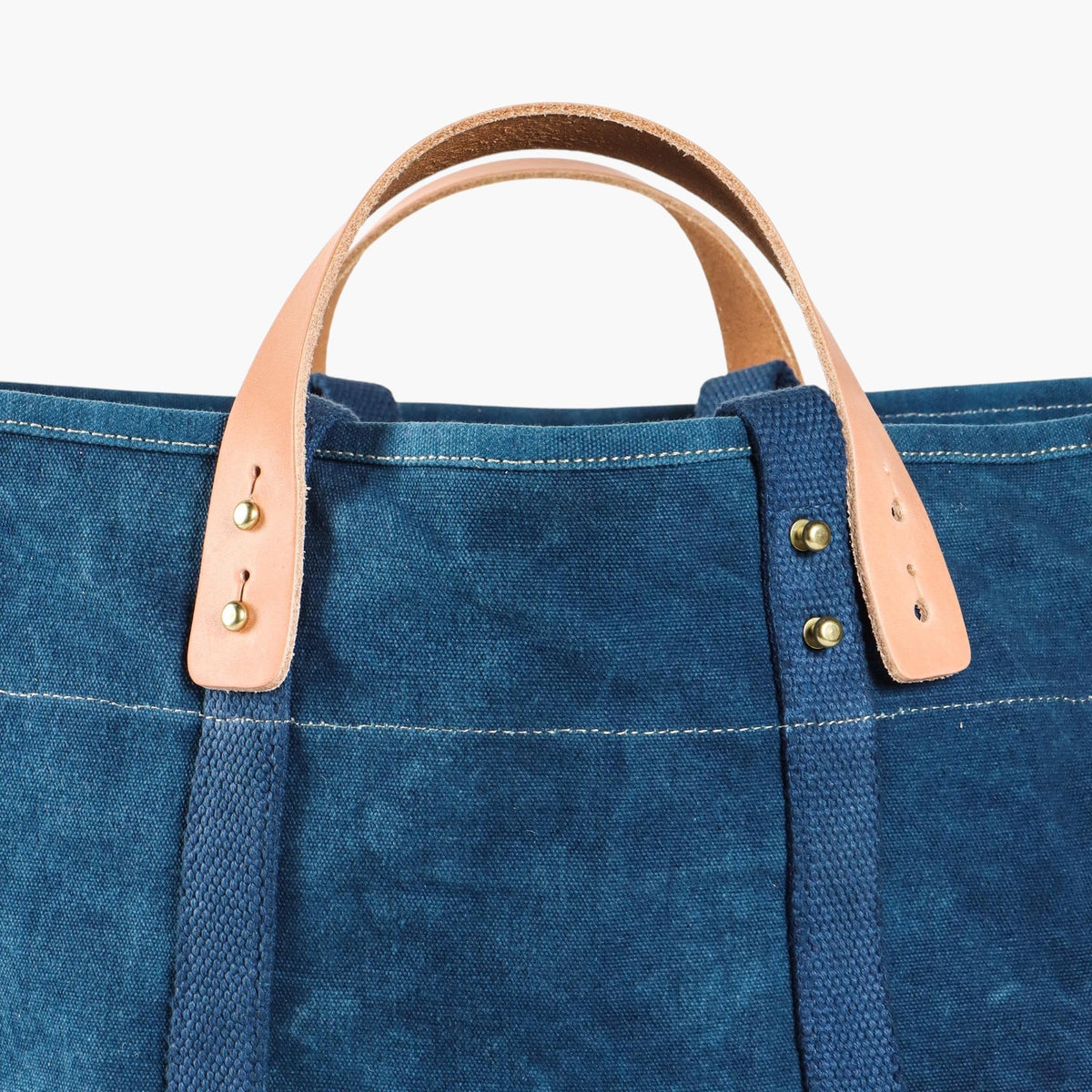 Close-up of the indigo tote's detachable natural leather handles, metal buttons, and blue webbing straps.
