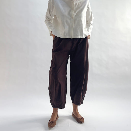 Full front view of model wearing Eleven Stitch chicory snap doreen pant with hands in pockets, showing elastic waistband, wide balloon leg tapering to snap hem at ankle, and deep espresso brown color, styled with cream button front shirt and taupe ballet flats