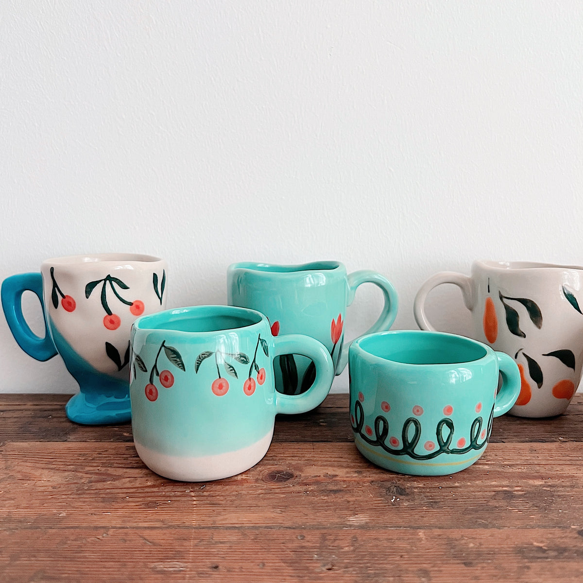 Close-up of turquoise mugs with cherry, tulip, and looping patterns beside cream fruit-patterned mugs, including a blue-footed cherry mug.
