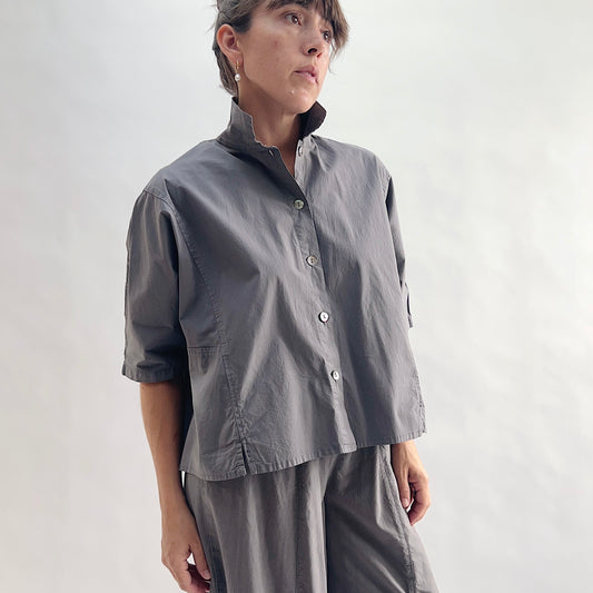 Three-quarter front view of model wearing Eleven Stitch eiffel tower camp shirt looking up, showing point collar, dark shell buttons, short sleeves with rolled cuffs, slightly cropped boxy fit, and deep slate grey color, styled with matching eiffel tower one pocket pant