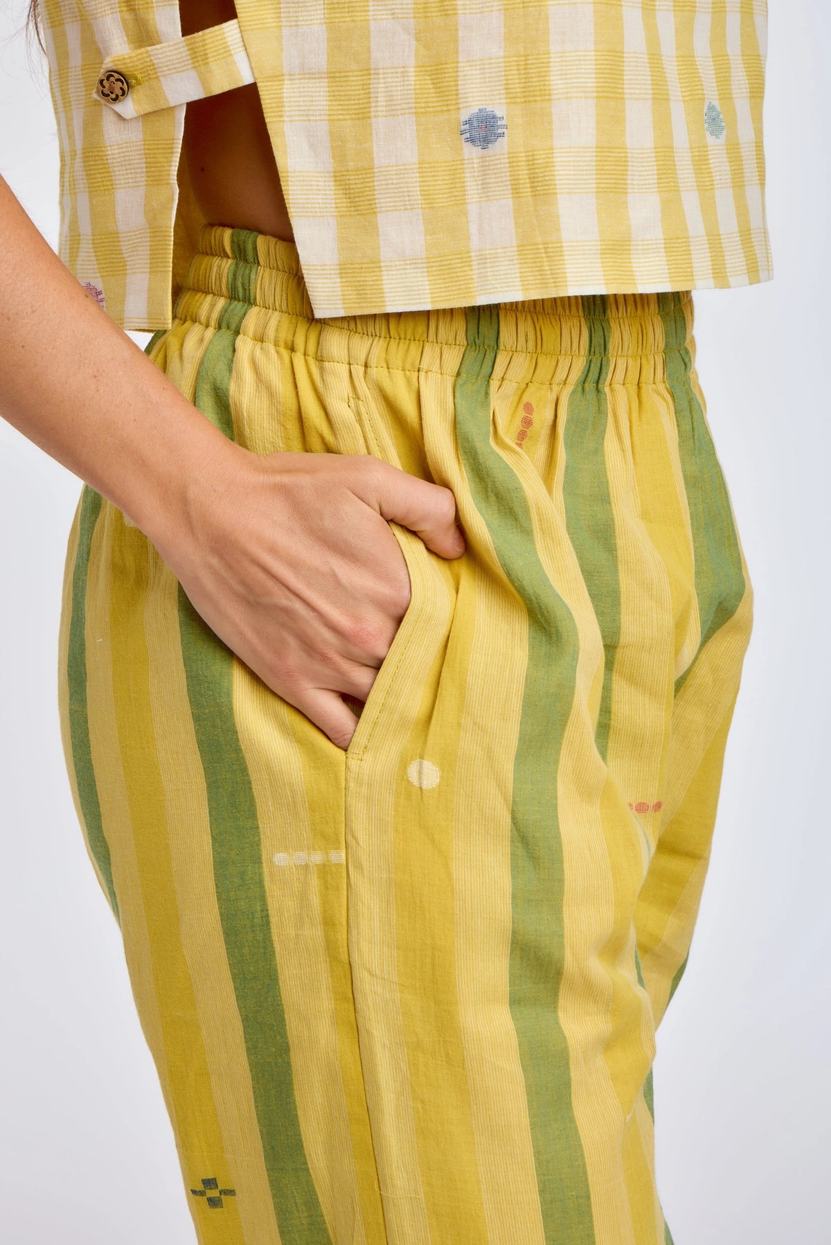 Detail view of DVAA Celeste Pant in Goldenrod waistband and side pocket, showing goldenrod yellow and sage green wide vertical stripe handwoven cotton with scattered small woven ikat motifs, wide elasticated waistband, and hand tucked into side pocket, styled with yellow handwoven check crop top
