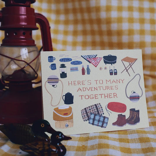 Small Adventure Adventures Together Card, illustrated camping gear surrounding the words “Here's to many adventures together,” displayed beside a red lantern.
