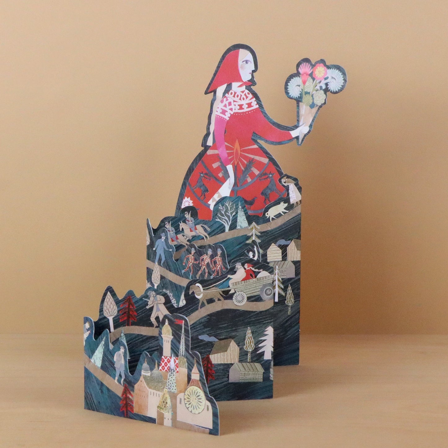 Storyteller die-cut concertina card by Sarah Young, front view fully extended to reveal a woman in a red dress holding a bouquet above layered dark teal concertina panels with fairy tale scenes including dancing skeletons, a horse-drawn cart, and a village with a church