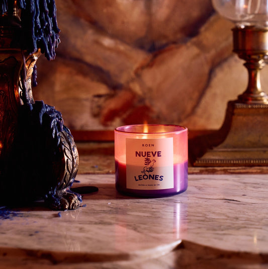 A Roen candle with a label 'NUEVE LEONES' burning inside a purple jar on a marble surface, with a richly colored artwork in the background.