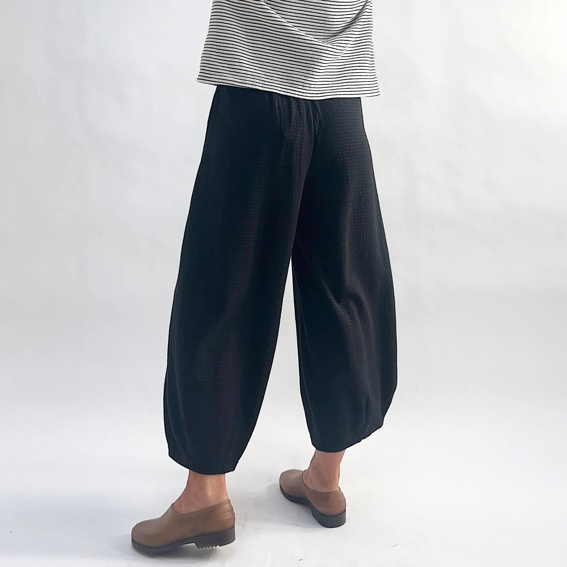 Three-quarter back view of model wearing black crinkle-texture cropped wide-leg pants, showing the elastic waist, wide rounded volume, and tapered ankle, styled with a white and black stripe long-sleeve top and taupe slip-on shoes against a white background.
