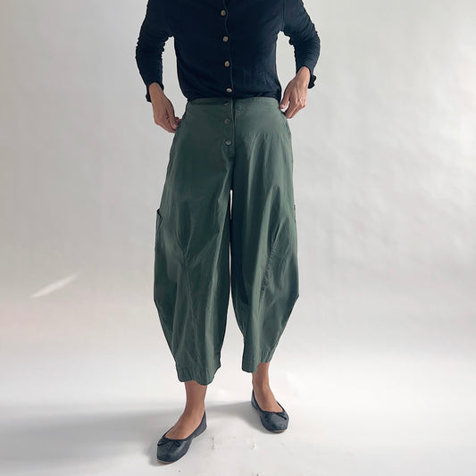 Full front view of model wearing Eleven Stitch konbu snap front pant with hands at waist, showing snap front waistband, wide balloon leg, large patch pockets, gathered elastic hem, and deep forest green color, styled with black long sleeve button front cardigan and black ballet flats