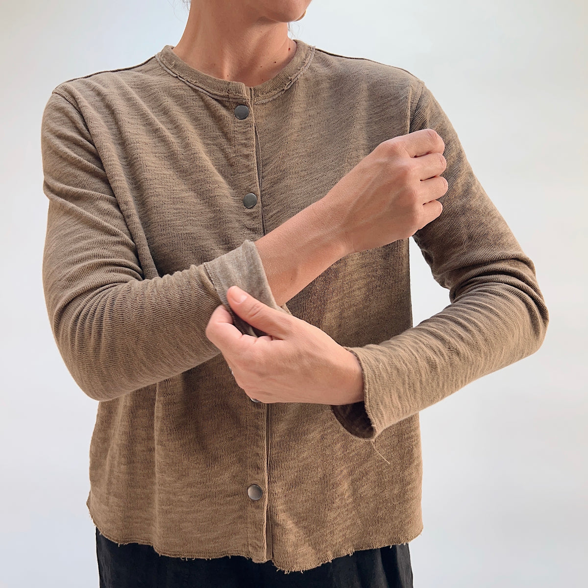 Close-up of model adjusting the sleeve of the Cut Loose Snap Front Cardigan in Winter Olive, showing snap button closure and raw hem detail in 100% cotton
