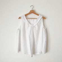 Load image into Gallery viewer, A white linen sleeveless top with a round neckline, hanging on a wooden hanger against a white wall.
