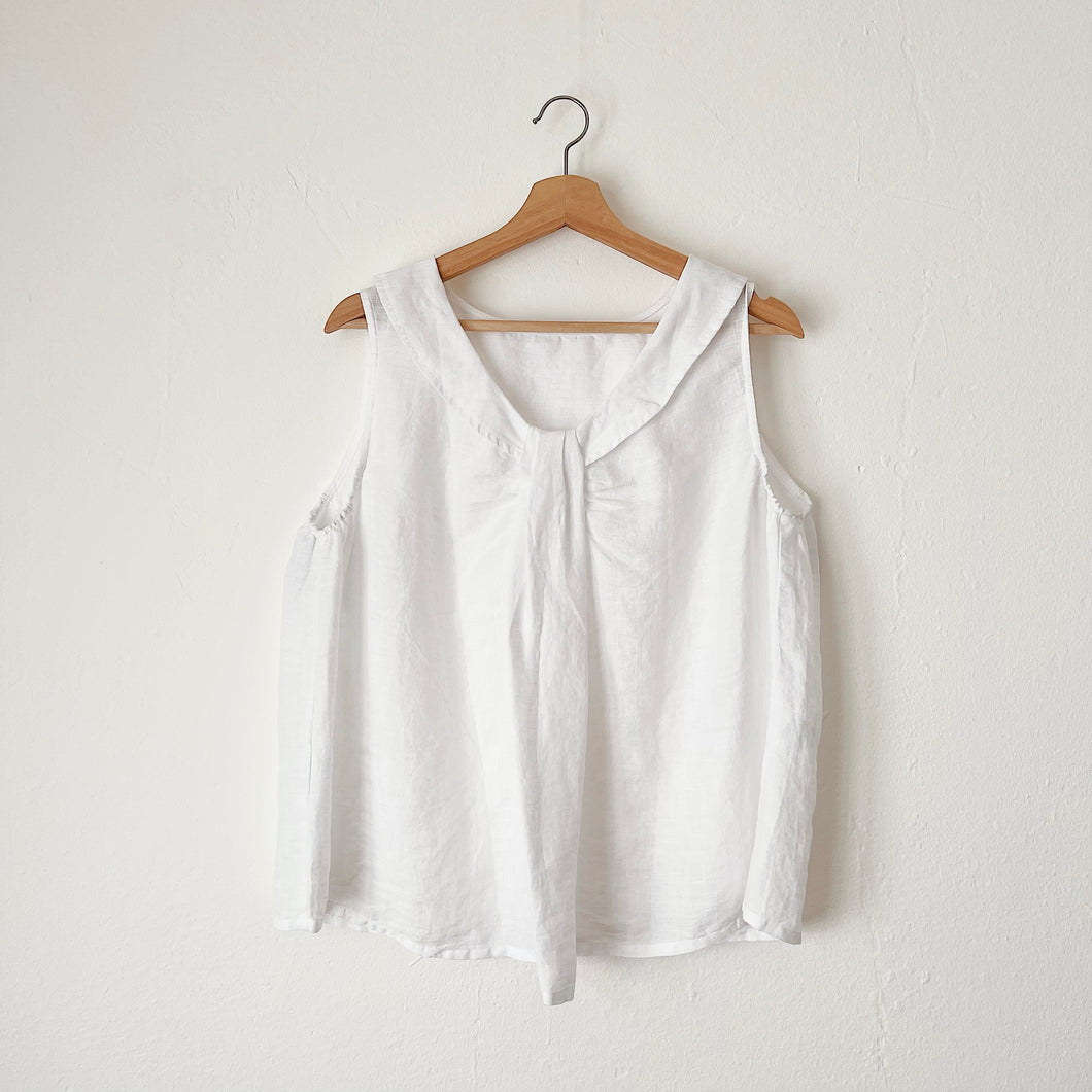 A white linen sleeveless top with a round neckline, hanging on a wooden hanger against a white wall.