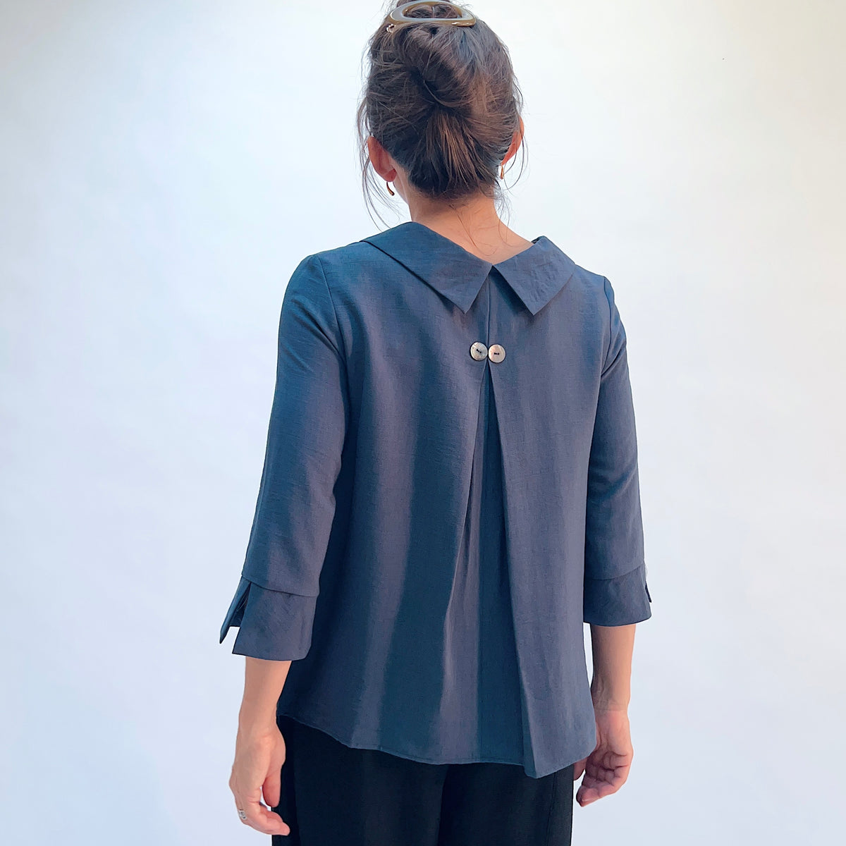 A woman viewed from behind wearing a slate blue-gray three-quarter sleeve blouse with a collar and two decorative buttons at the back, paired with black pants, against a plain light background.