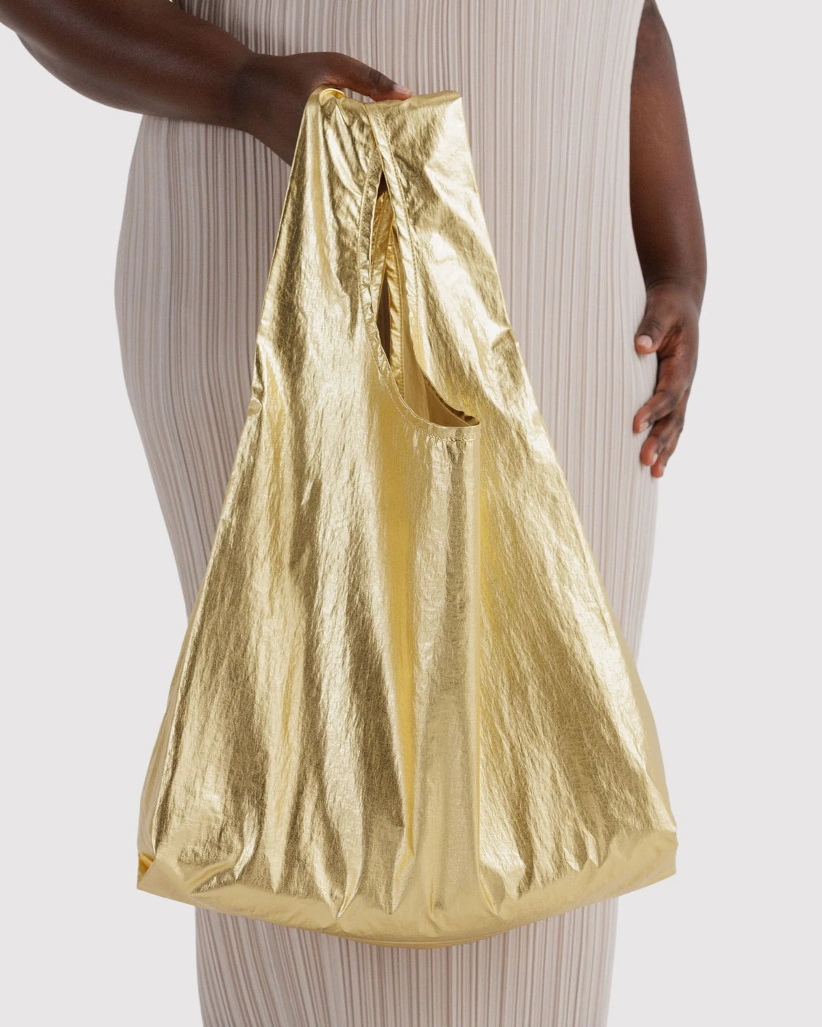BAGGU Standard Baggu in Metallic Gold, held at a model’s side to show the bag’s size and reflective gold finish.
