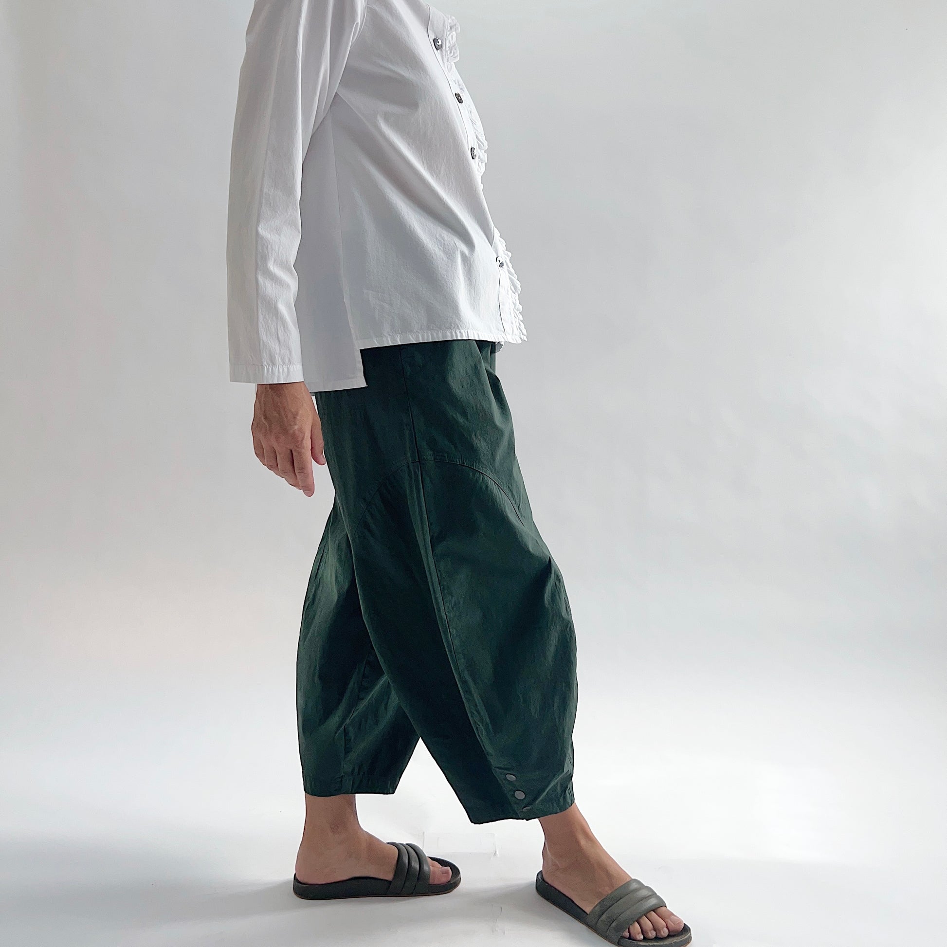 Side view of model wearing Eleven Stitch konbu snap doreen pant in motion, showing elastic waistband, wide balloon leg tapering to snap hem at ankle, side seam pocket, and deep forest green color, styled with white blossom collar shirt and dark slide sandals