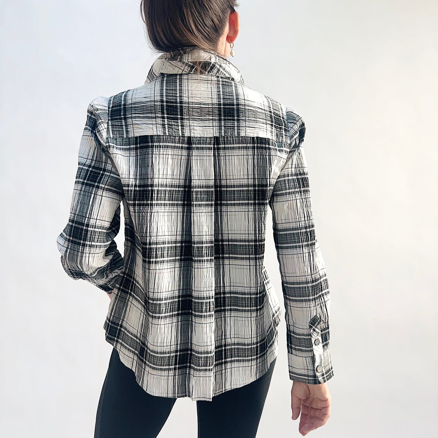 Back view of model wearing a white and charcoal windowpane plaid crinkle pullover shirt, showing the back yoke with center pleat, long sleeves with buttoned cuffs, and relaxed A-line hem, styled with black slim pants against a white background.
