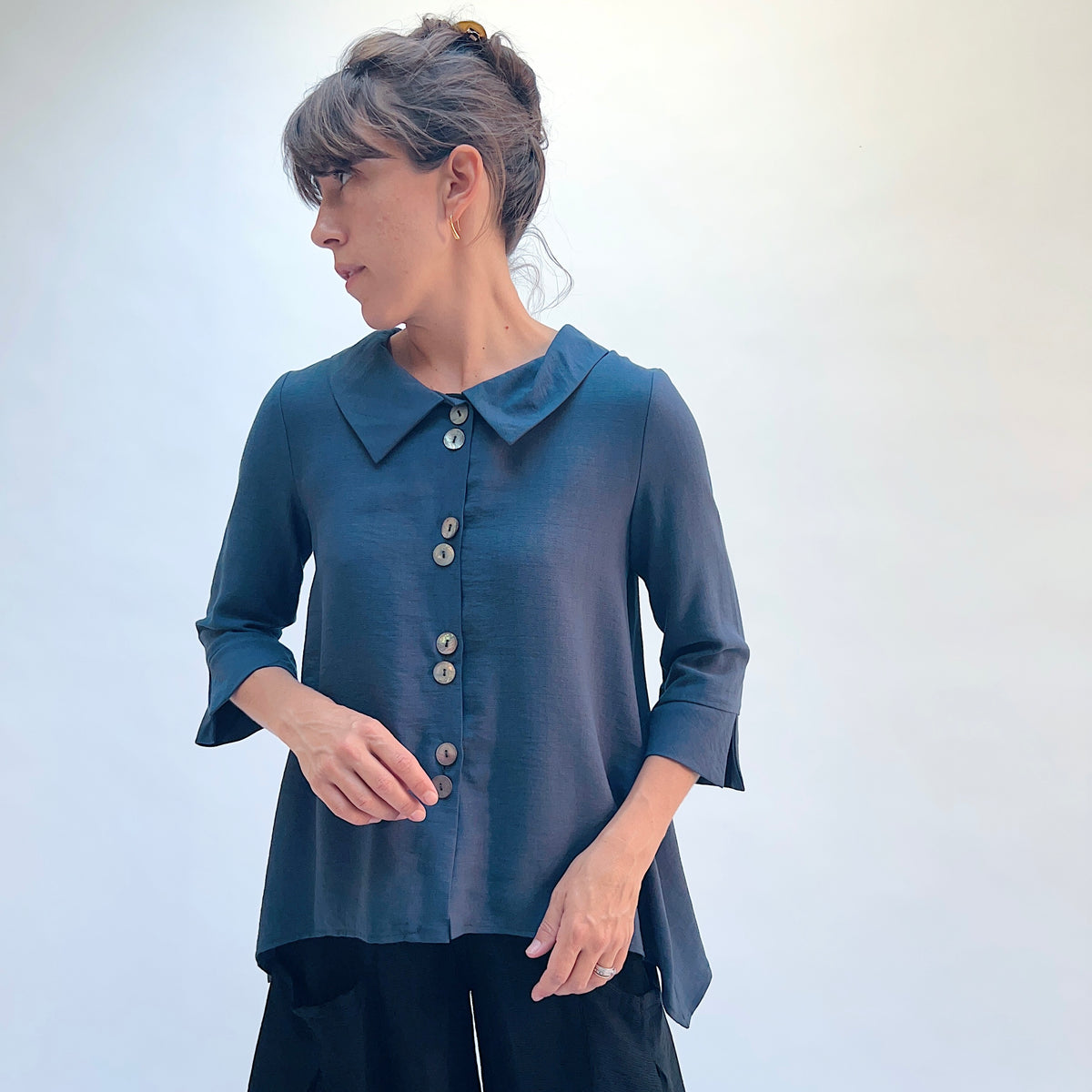 A woman wearing a slate blue button-down top with a pointed collar and black pants, standing against a light gray background.