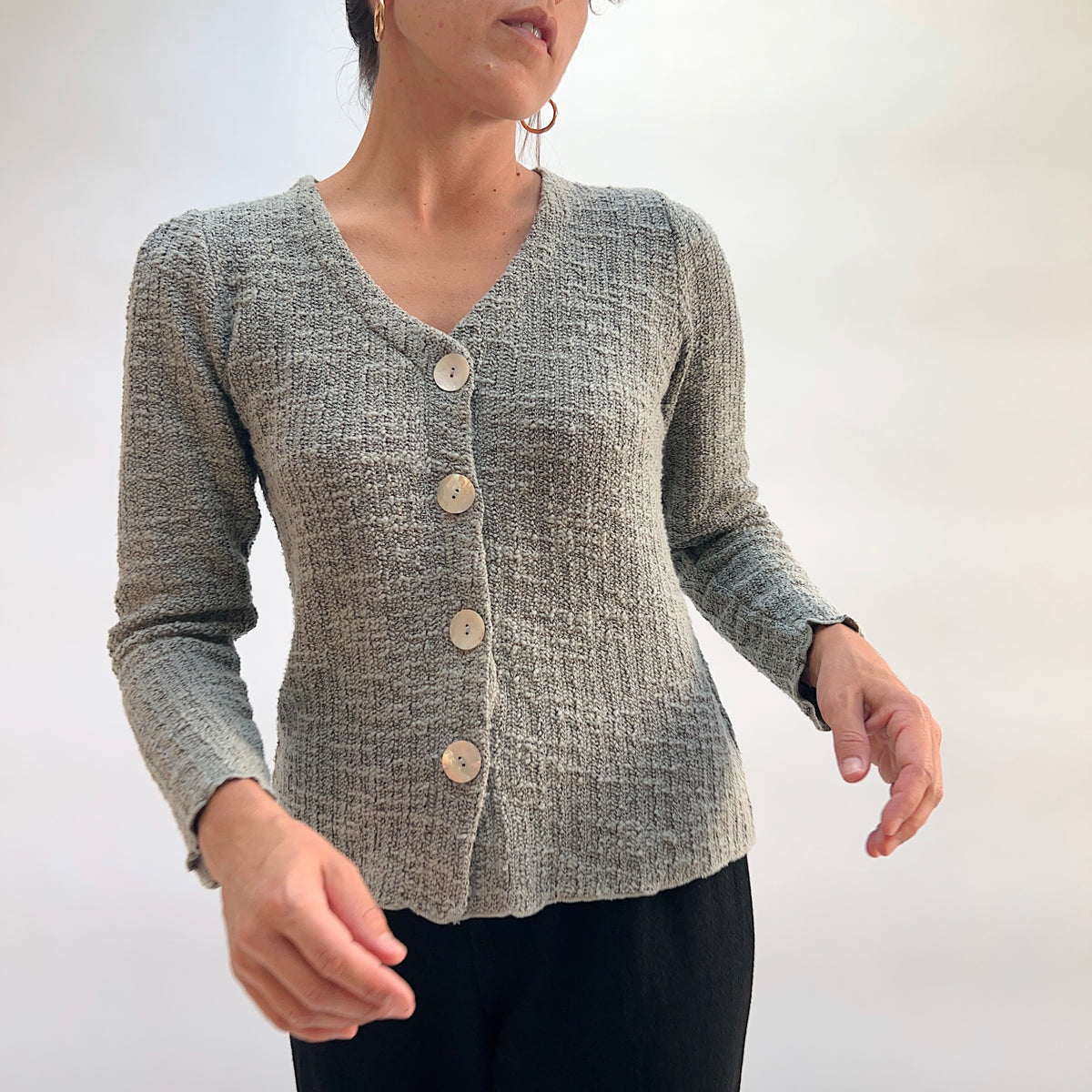 Model wearing the Cut Loose Puff Sleeve Cardigan in Squirrel, back view with hands behind back, showing gently puffed shoulder seam, ribbed back panel, and scalloped lettuce hem of warm grey-taupe marled knit cardigan, styled with black pants
