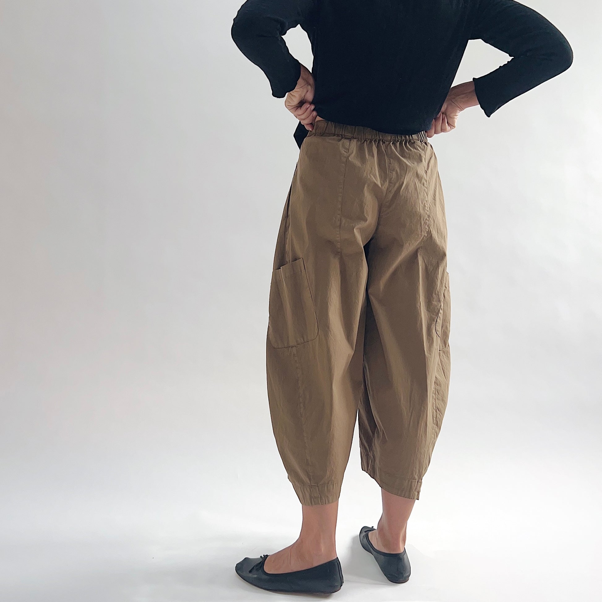 Back view of model wearing Eleven Stitch winter olive snap front pant with hands on waist, showing elastic back waistband, wide balloon leg, large back patch pocket, gathered elastic ankle hem, and warm olive tan color, styled with black button front top and black ballet flats