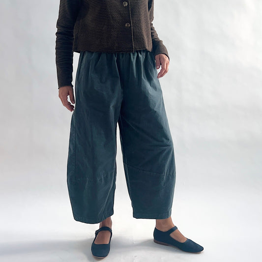 Full front view of model wearing iron blue-gray mini cord lantern pants, arms at sides, showing the gathered elastic waist, full rounded silhouette, and tapered ankle seam detail, styled with the matching chocolate striped cropped cardigan and blue mary jane flats against a white background.
