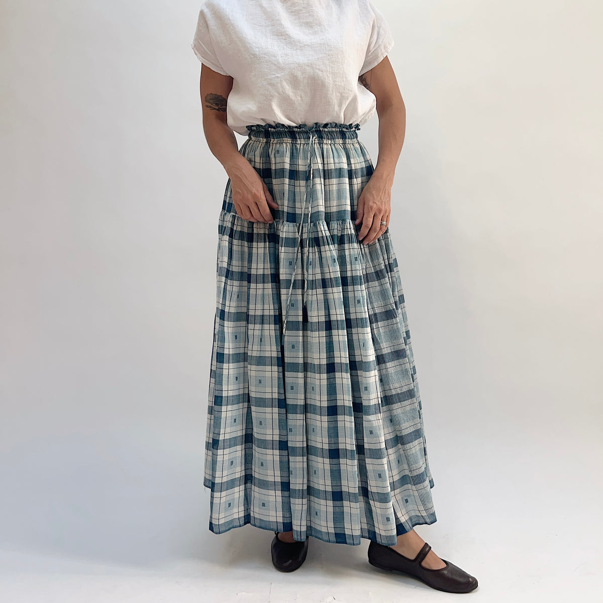 DVAA Denise Skirt in Blue Plaid, full-length tiered maxi skirt with ruffled elastic waistband, styled with a white tee and black Mary Jane flats — front view
