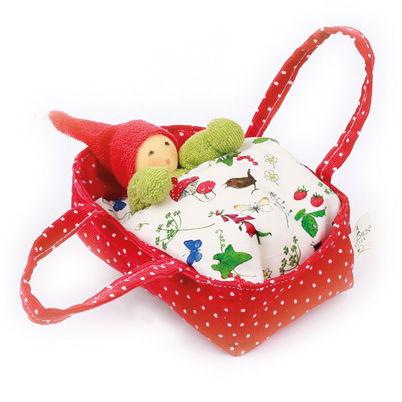 An organic cotton Waldorf baby doll with a hand-painted face, positioned in a red flower bed with a multicolored blanket and a green knitted hat.