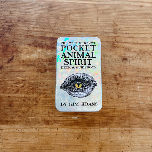 The Wild Unknown Pocket Animal Spirit Deck and Guidebook by Kim Krans, top view of closed holographic iridescent tin box on a wood surface, featuring a detailed hand-drawn reptilian eye illustration and the title in black serif lettering
