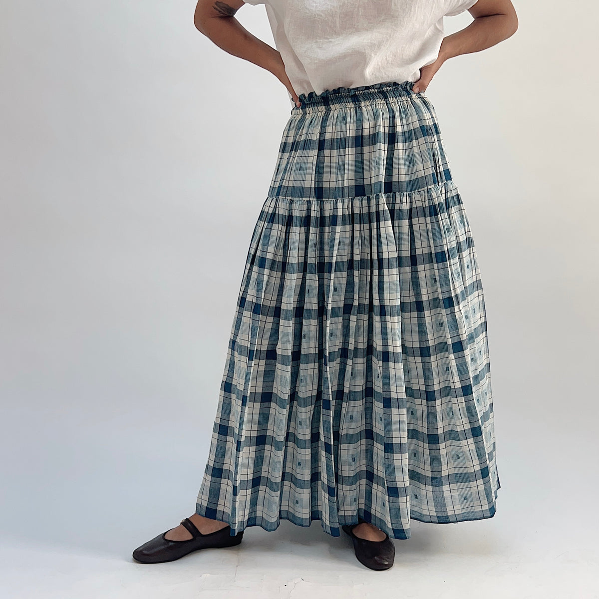 DVAA Denise Skirt in Blue Plaid, tiered cotton maxi skirt with elastic waistband, styled with a white tee and black Mary Jane flats — straight-on front view
