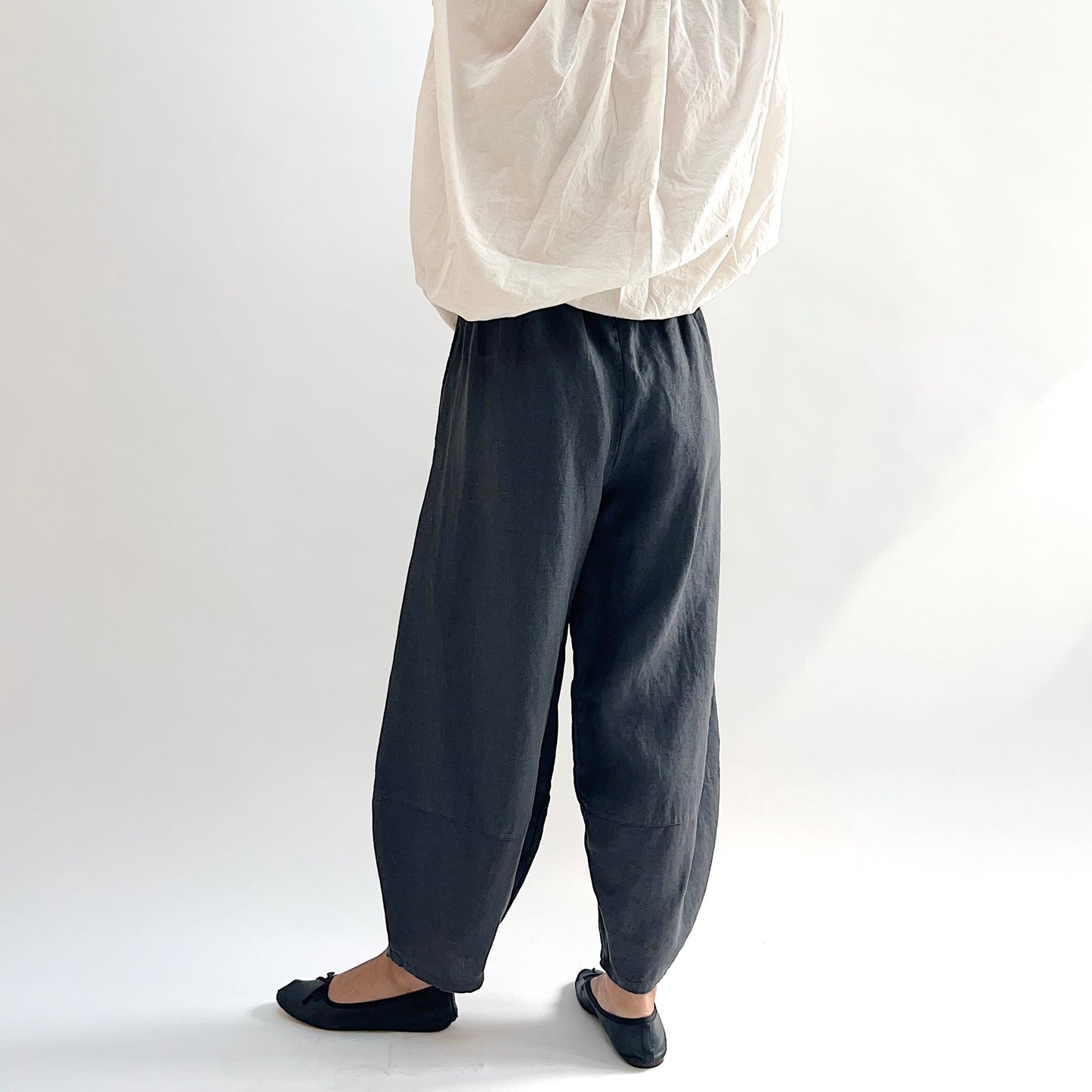 Back view of model wearing the Bryn Walker Heavy Linen Oliver Pant in Moonlight, showing elastic waistband, full barrel-leg drape, and tapered ankle in deep moonlight charcoal heavy linen, styled with cream oversized top and black ballet flats
