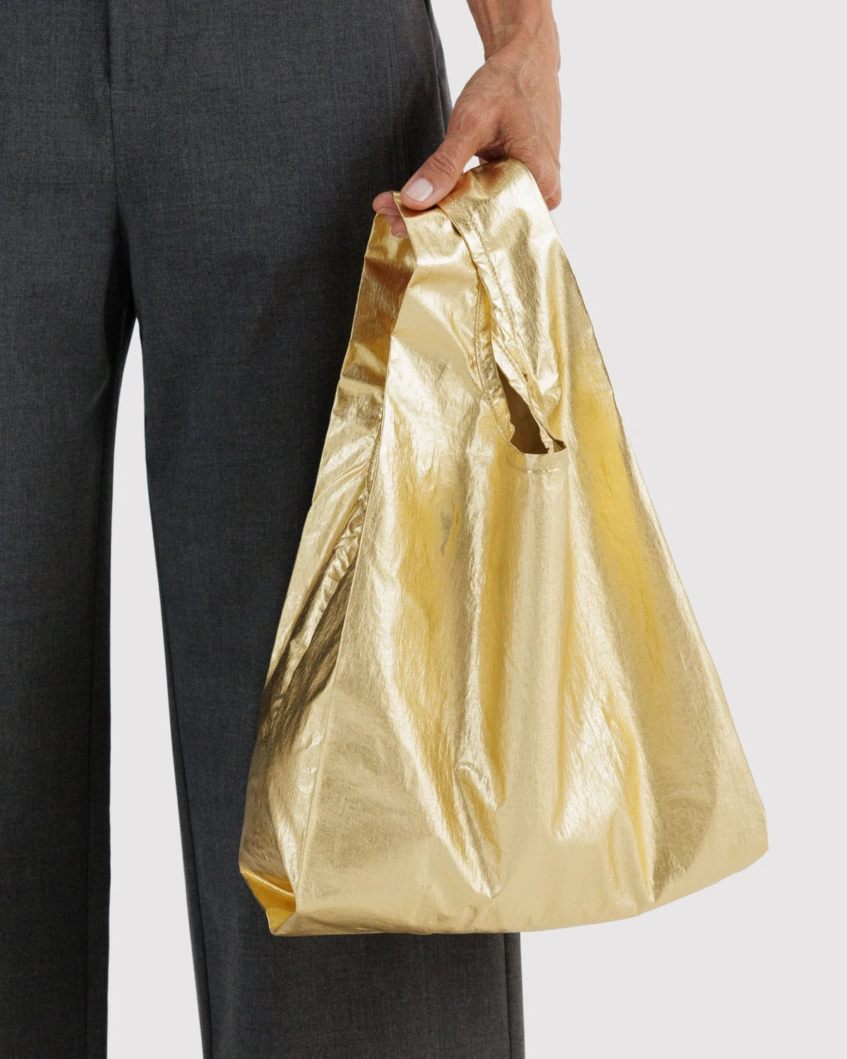 BAGGU Baby Baggu in Metallic Gold, held at a model’s side to show the small bag’s size and reflective gold finish.
