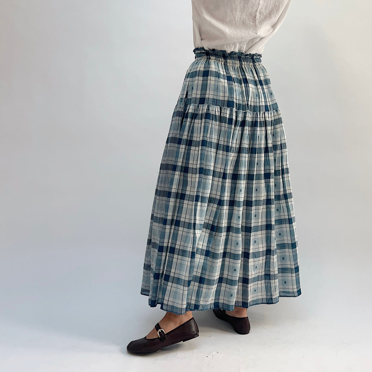 DVAA Denise Skirt in Blue Plaid, tiered maxi skirt with ruffled elastic waist, styled with a white tee — back three-quarter view with arms raised

