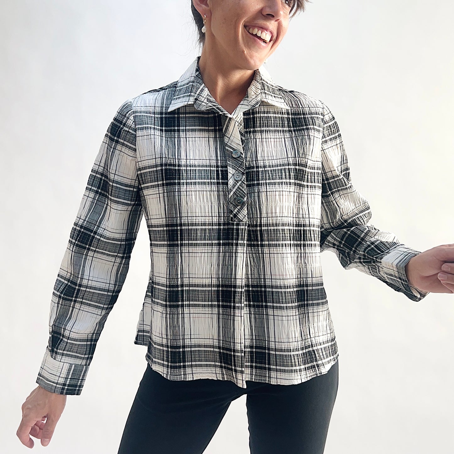 Full front view of smiling model wearing a white and charcoal windowpane plaid crinkle pullover shirt, arms in motion, showing the point collar, half placket with matte buttons, and relaxed A-line hem, styled with black slim pants against a white background.
