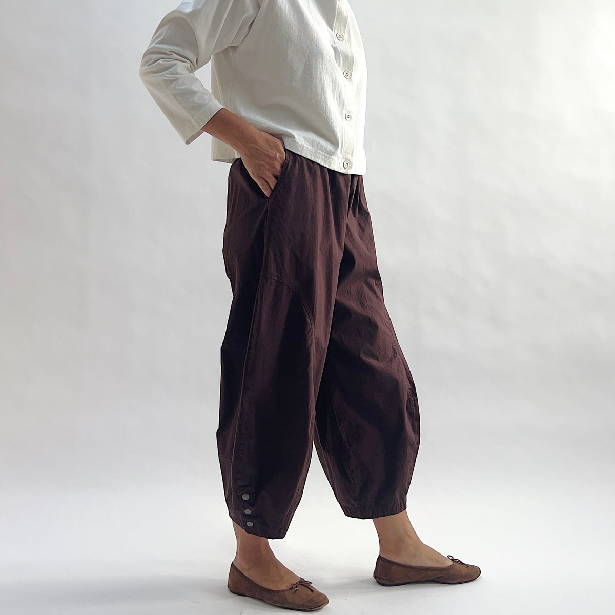 Three-quarter front view of model wearing Eleven Stitch chicory snap doreen pant with hand in pocket, showing elastic waistband, wide balloon leg tapering to snap hem at ankle, side seam pocket, and deep espresso brown color, styled with cream button front shirt and taupe ballet flats


