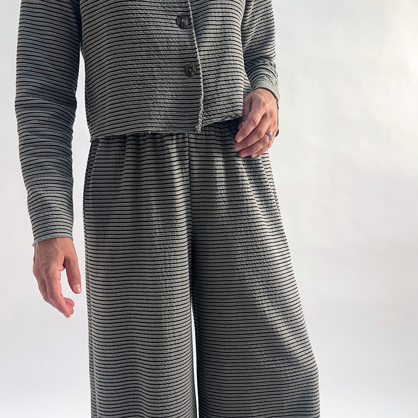 Close crop of model wearing squirrel and black fine horizontal stripe wide-leg cropped pants with dart shaping, showing the elastic waist and crinkle-texture fabric, styled with the matching striped asymmetrical cardigan against a white background.
