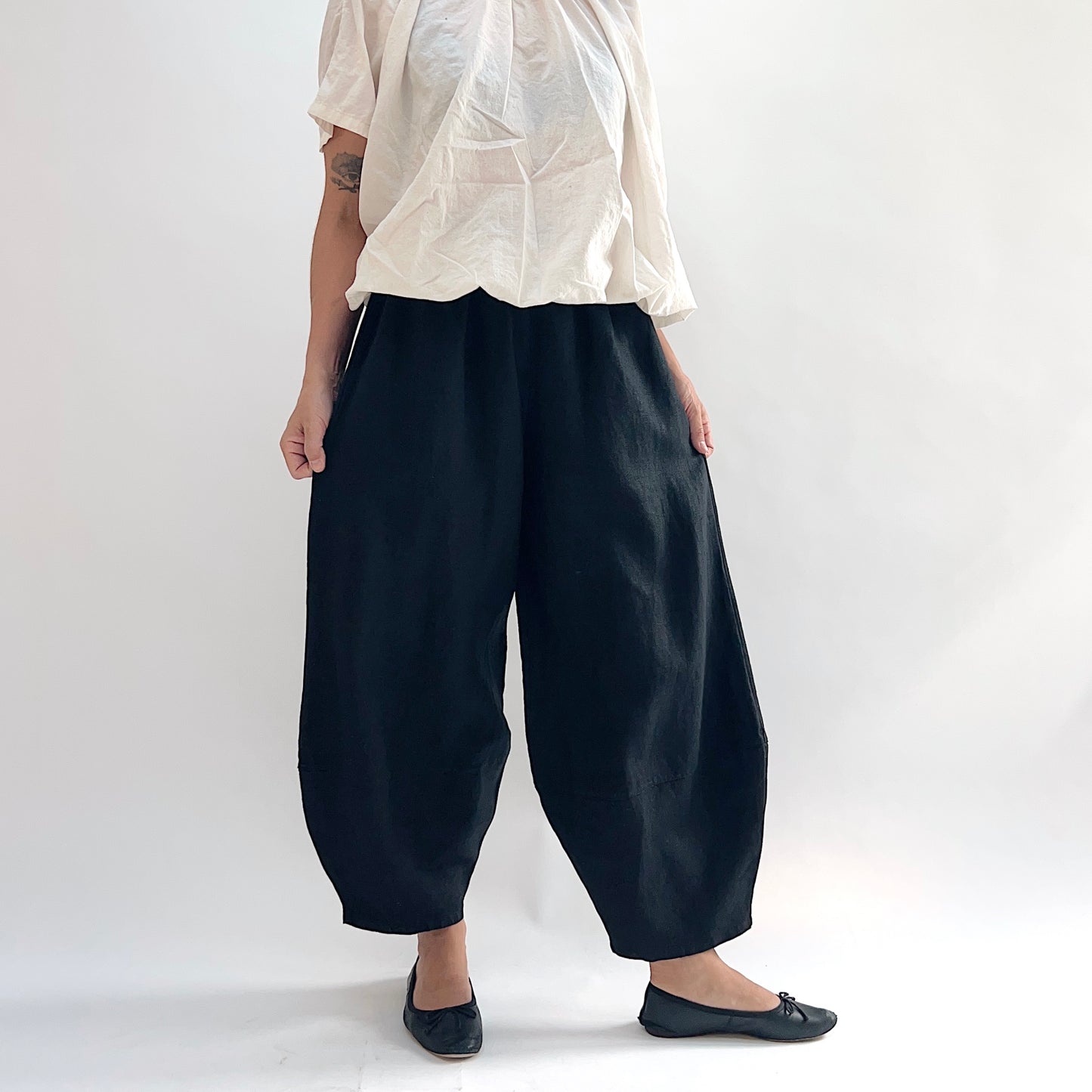 Model wearing the Bryn Walker Heavy Linen Oliver Pant in Black, front view showing barrel-leg silhouette, elastic waistband, side pockets, and tapered ankle in black heavy linen, styled with cream short-sleeve linen top and black ballet flats
