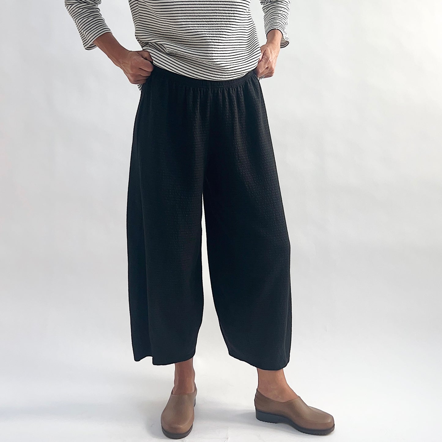 Full front view of model wearing black crinkle-texture cropped wide-leg pants with hands at elastic waist, showing the wide rounded silhouette and tapered ankle, styled with a white and black stripe long-sleeve top and taupe slip-on shoes against a white background.
