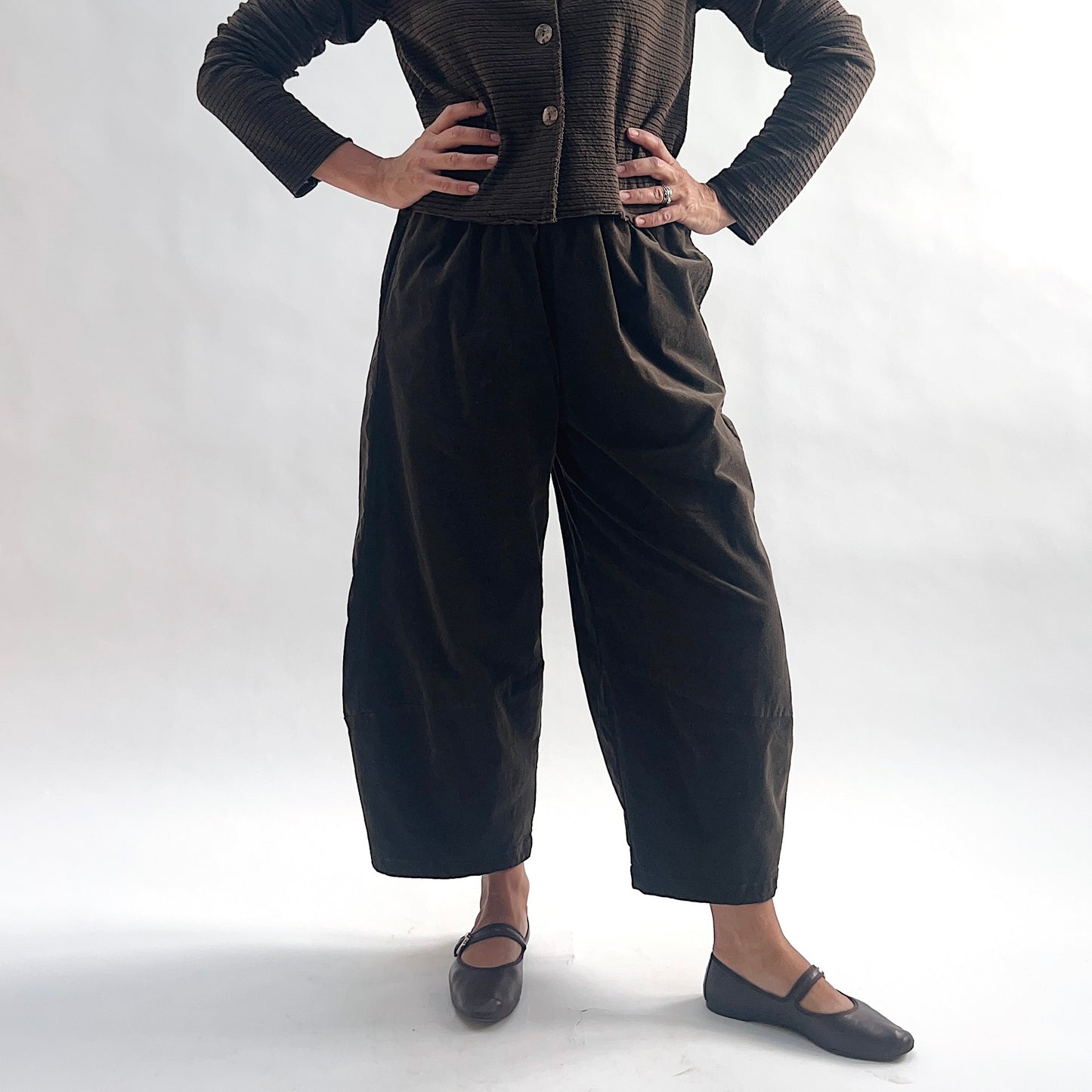 Close front view of model wearing chocolate mini cord lantern pants with elastic gathered waist and tapered ankle, hands at waist, showing the rounded lantern silhouette, styled with the matching striped cropped cardigan and dark mary jane flats against a white background.
