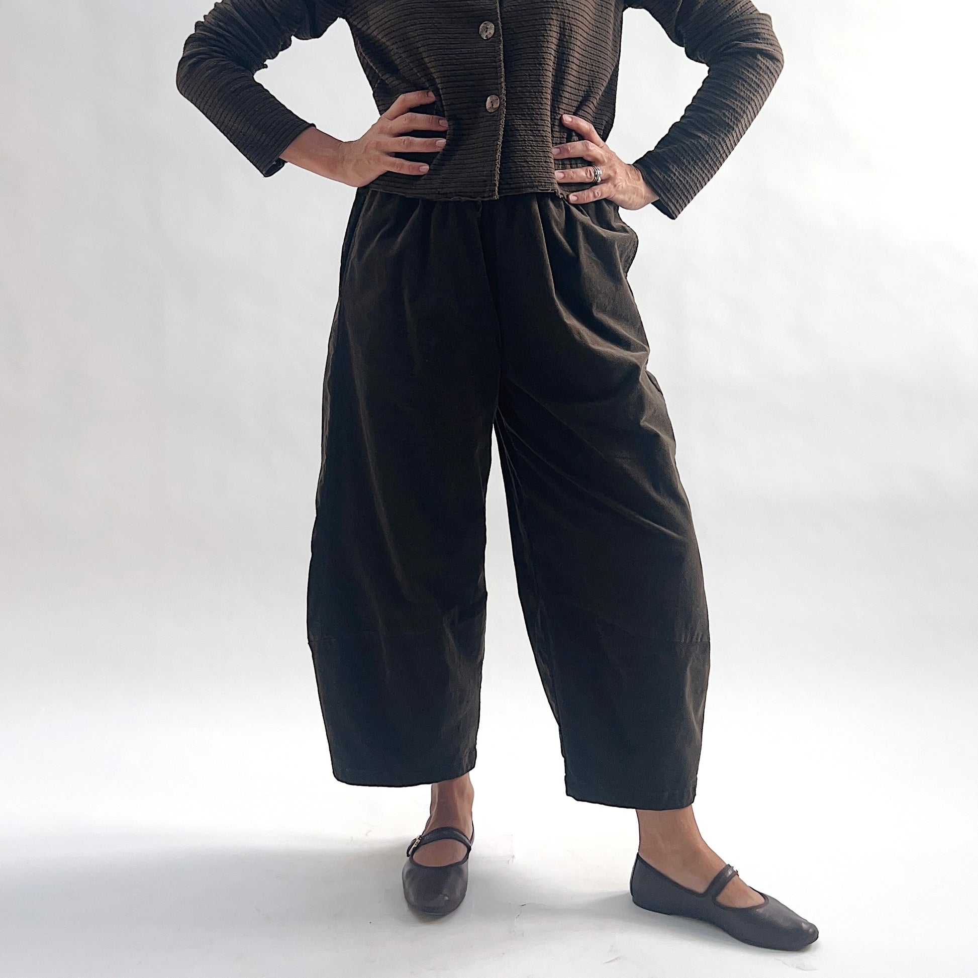Close front view of model wearing chocolate mini cord lantern pants with elastic gathered waist and tapered ankle, hands at waist, showing the rounded lantern silhouette, styled with the matching striped cropped cardigan and dark mary jane flats against a white background.
