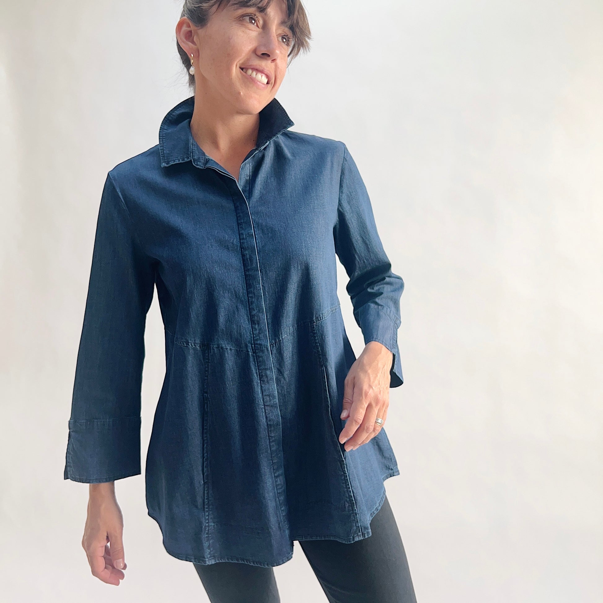 Full front view of model wearing a lightweight dark indigo denim tunic shirt with hidden front placket, point collar, and 3/4 sleeves with slit cuffs, showing the empire seam, flared A-line hem, and curved shirttail, styled with black slim pants against a white background.
