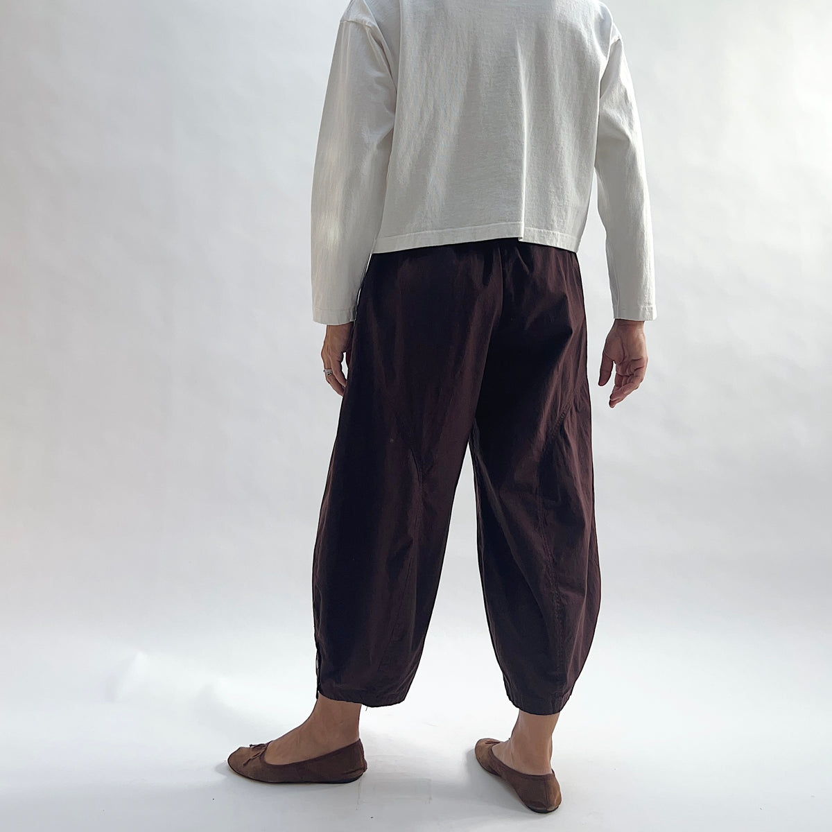 Back view of model wearing Eleven Stitch chicory snap doreen pant, showing elastic waistband, wide balloon leg tapering to snap hem at ankle, side seam detailing, and deep espresso brown color, styled with cream long sleeve top and taupe ballet flats