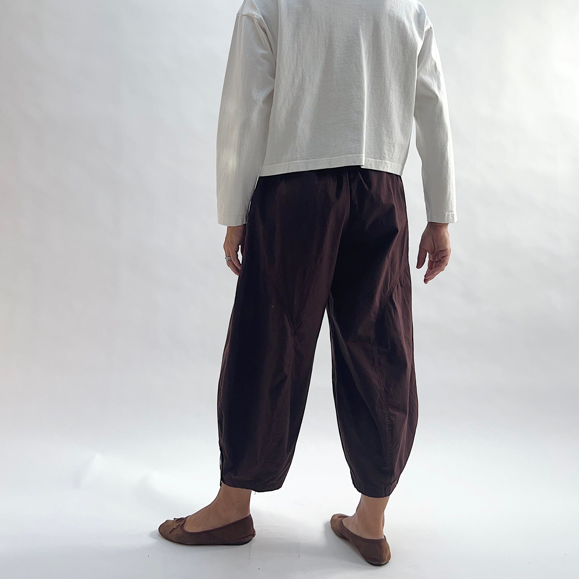 Back view of model wearing Eleven Stitch chicory snap doreen pant, showing elastic waistband, wide balloon leg tapering to snap hem at ankle, side seam detailing, and deep espresso brown color, styled with cream long sleeve top and taupe ballet flats