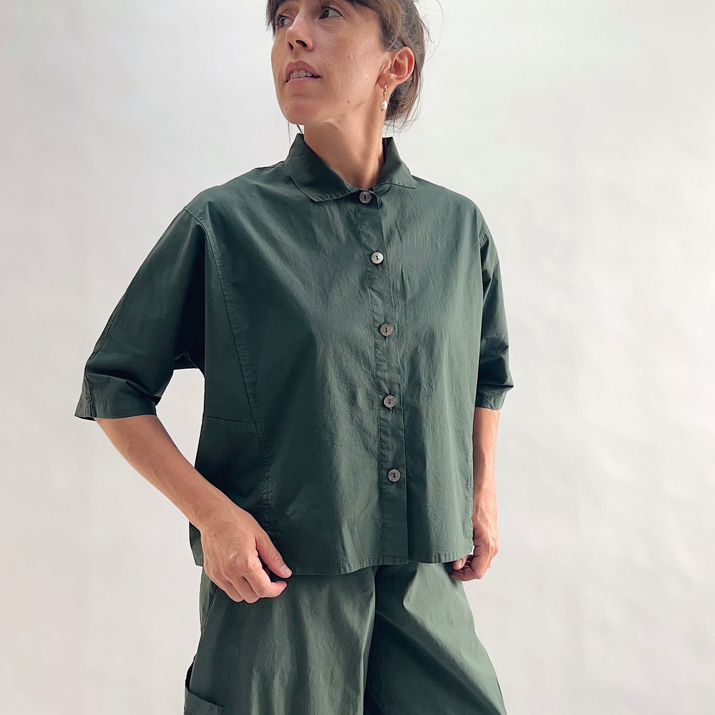 Close front view of model wearing Eleven Stitch konbu camp shirt looking up, showing rounded collar, dark shell buttons, short sleeves, slightly cropped boxy fit, and deep forest green color, styled with matching konbu snap front pant