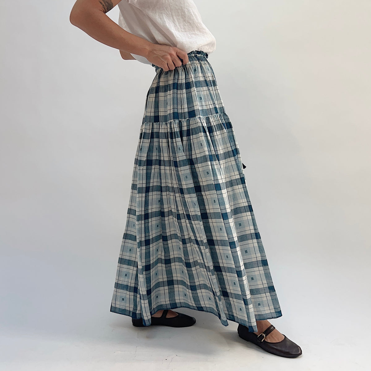 DVAA Denise Skirt in Blue Plaid, tiered maxi skirt with ruffled elastic waist and tassel tie detail, styled with a white tee — side view showing full length and drape
