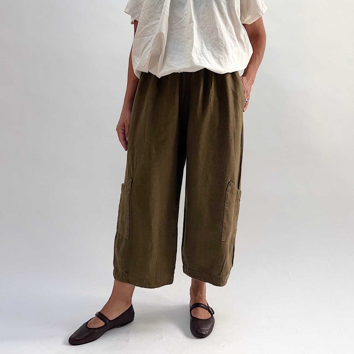 Model wearing the Bryn Walker Heavy Linen Casbah Pant in Partridge, front view with hand in hip pocket, showing elastic waistband, patch pockets, and relaxed wide-leg silhouette in olive linen, with dark flats
