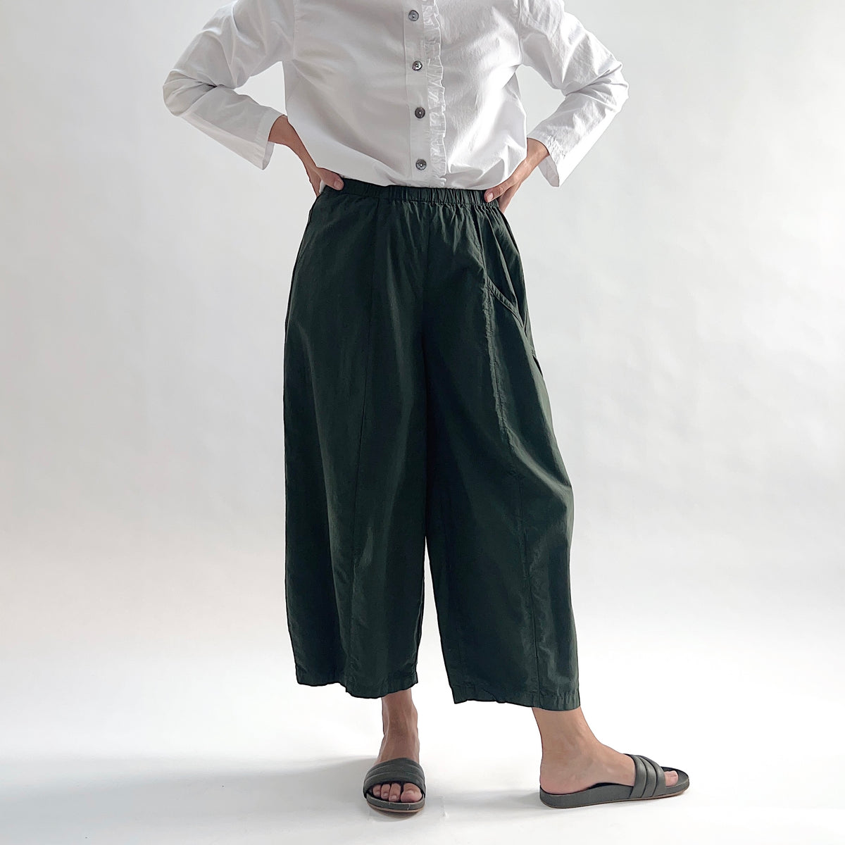 Full front view of model wearing Eleven Stitch konbu one pocket pant with hands on hips, showing elastic waistband, wide curved-seam cropped leg, large curved side pocket, and deep forest green color, styled with white long sleeve button front shirt and dark slide sandals