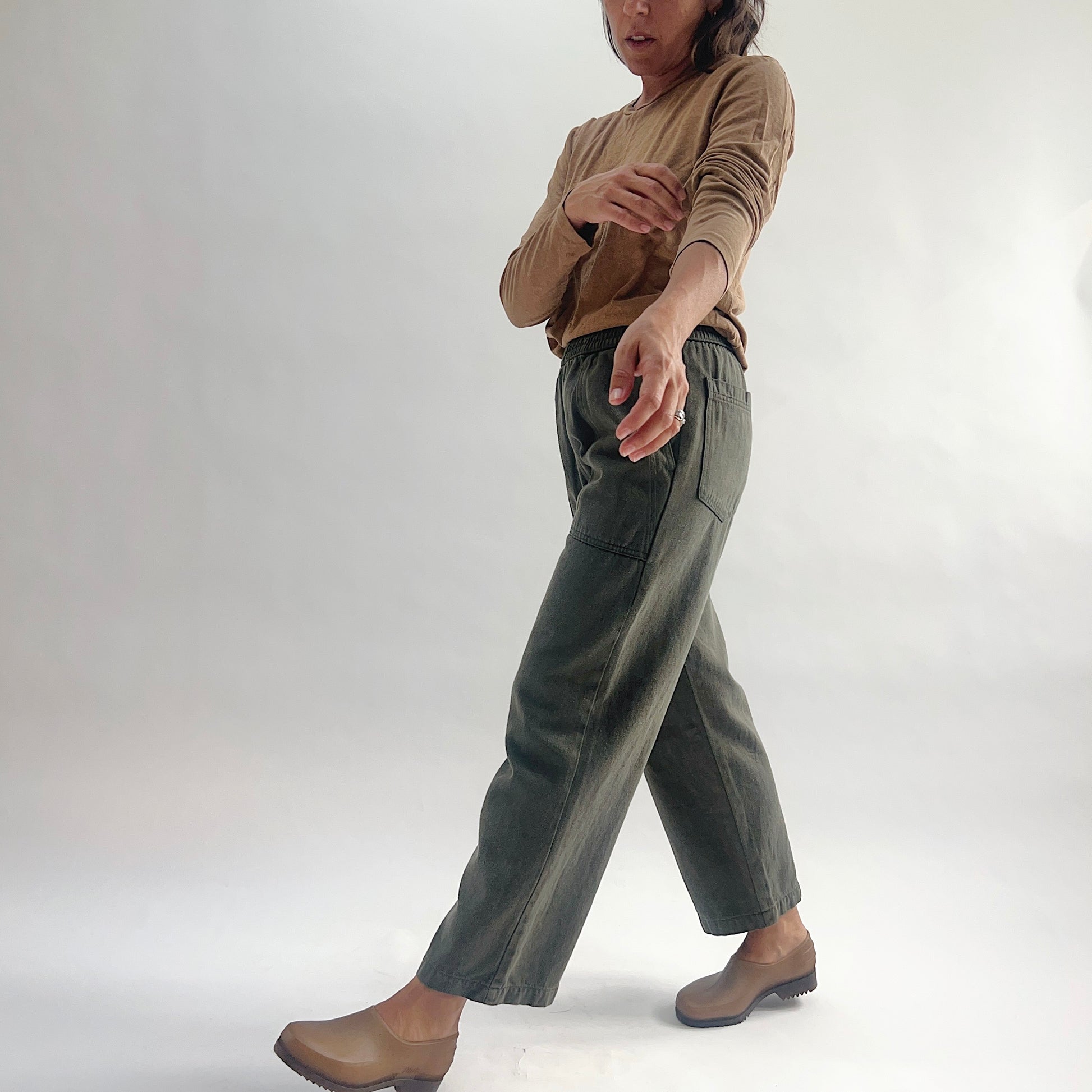 Side view of model mid-stride in olive green wide-leg pull-on pants with elastic waist and patch pocket, paired with a tan long-sleeve top and brown clogs.
