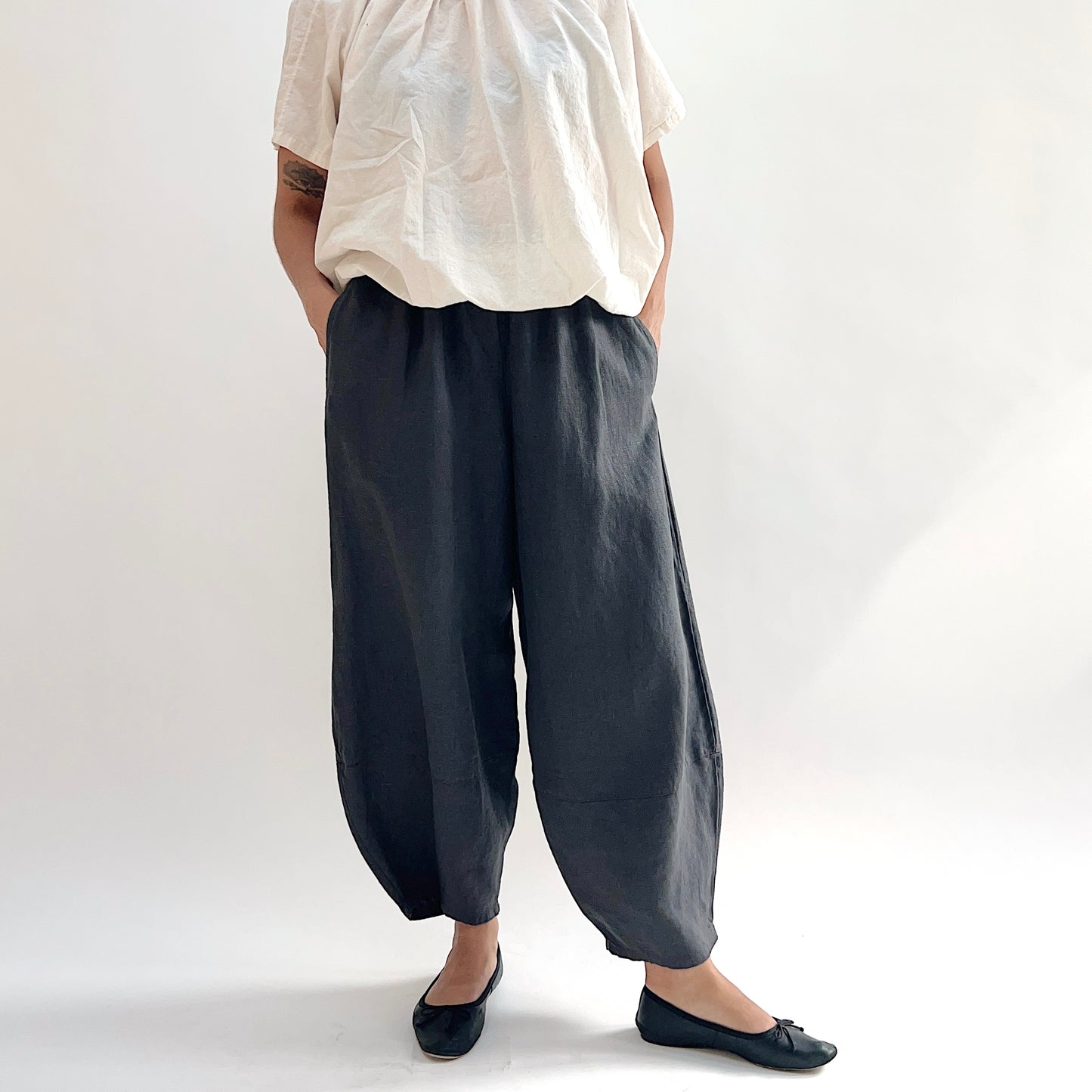 Model with hands in pockets wearing the Bryn Walker Heavy Linen Oliver Pant in Moonlight, front view showing barrel-leg silhouette, elastic waistband, side pockets, and tapered ankle in deep moonlight charcoal heavy linen, styled with cream short-sleeve top and black ballet flats
