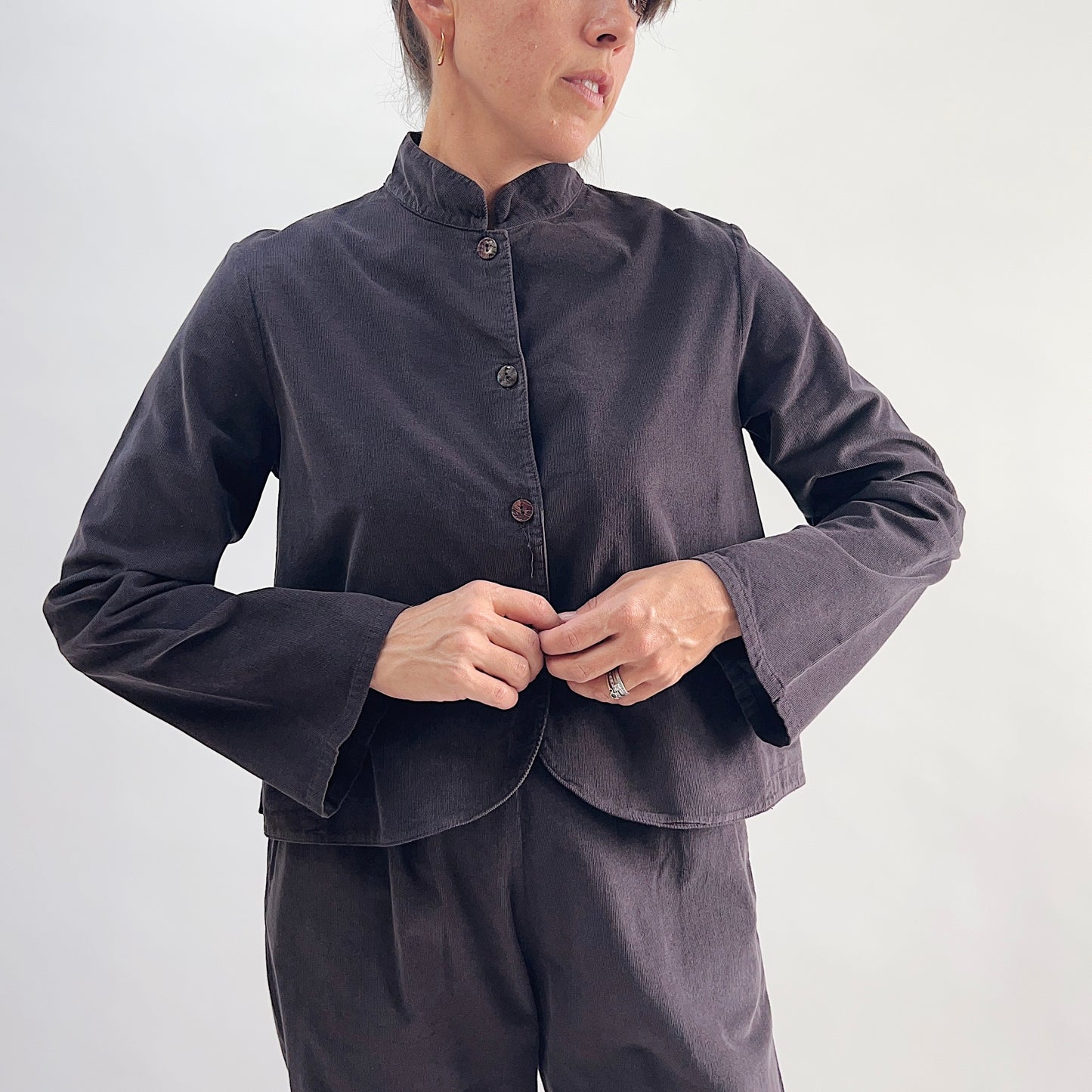 Model buttoning the Bryn Walker Corduroy Standing Collar Crop Jacket in Fig, close-up front view showing standing collar, mother of pearl buttons, wide sleeves with slit cuff detail, and deep fig pinwheel corduroy, styled with matching corduroy pants
