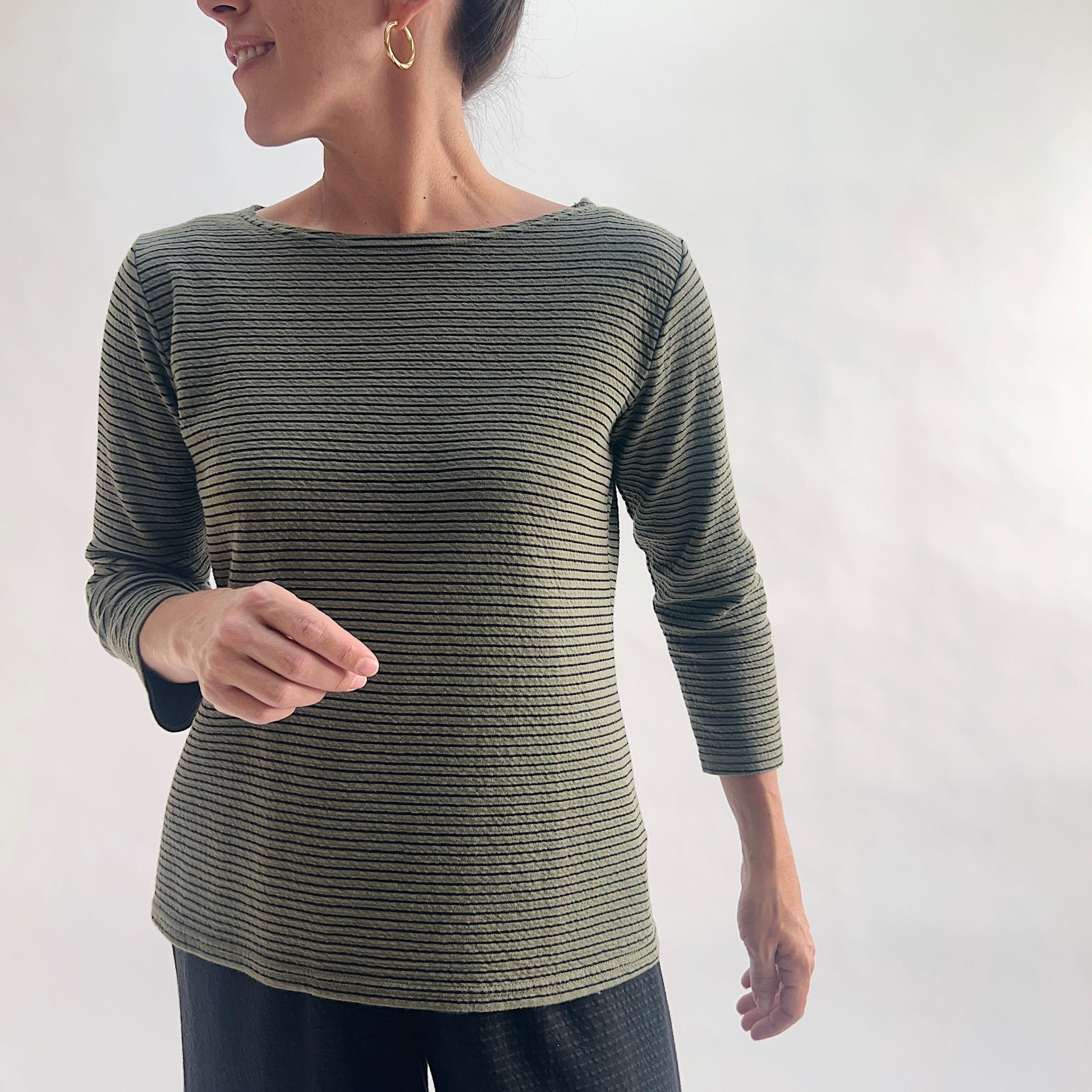 Smiling close front view of model wearing a kelp green tonal textured stripe boatneck tee with 3/4 sleeves, hand raised at side, showing the wide boatneck neckline and crinkle-texture fabric, styled with black wide-leg pants against a white background.
