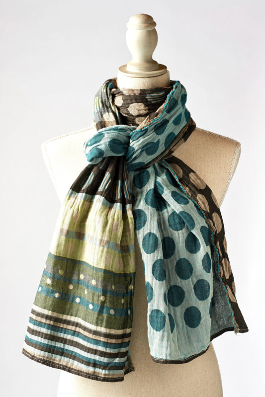 Létol Coline Scarf in Turquoise, draped and knotted on a linen dress form against a light grey background, showing a reversible Jacquard-woven organic cotton scarf — one side featuring large turquoise polka dots on a light blue ground, the other showing olive and taupe horizontal stripes with small dot accents and a bold black and teal striped border — with the woven fringe edge visible at the hem
