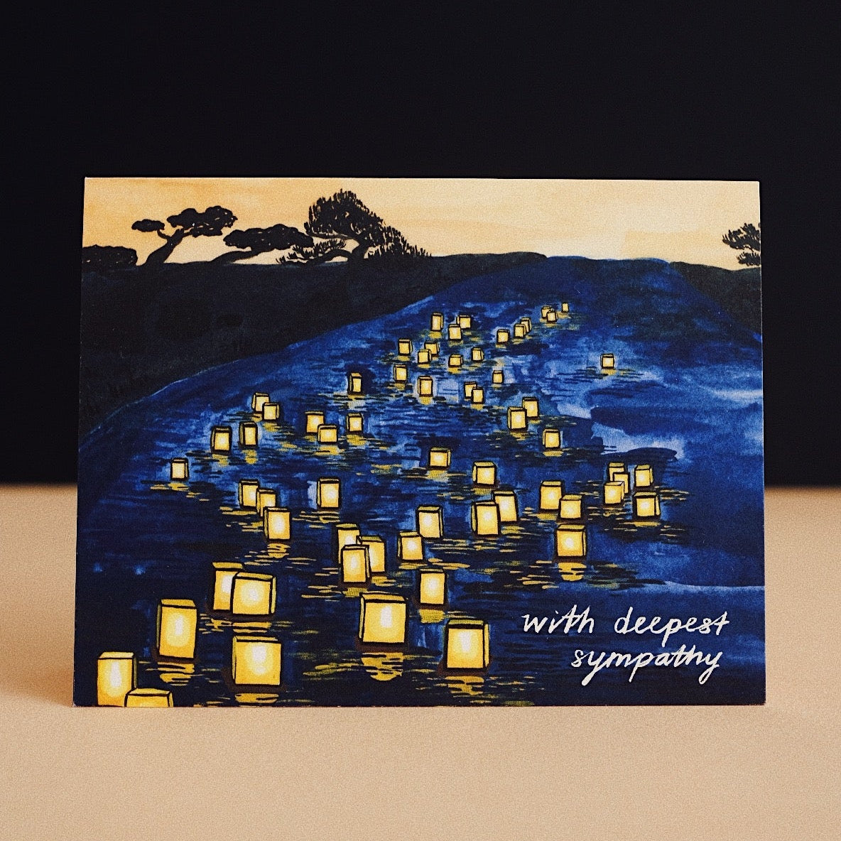 Small Adventure Japanese Lanterns Sympathy Card, glowing yellow lanterns floating on blue water beneath silhouetted trees, with the words “with deepest sympathy.”
